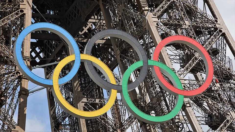 Five large interlocked rings in blue, yellow, black, green and red hanging on an iron lattice tower.