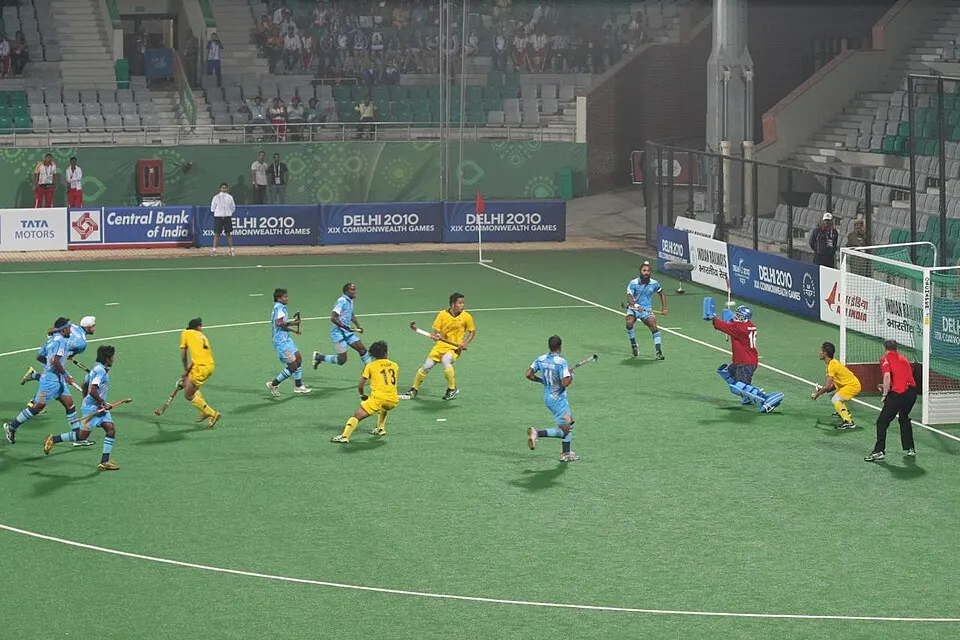 A field hockey match on green turf with players in sky blue and yellow kit near the goal.