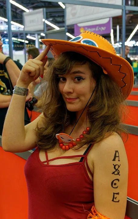 A smiling cosplayer in an orange cowboy hat with goggles on it, a red bead necklace and letters painted on the upper arm.