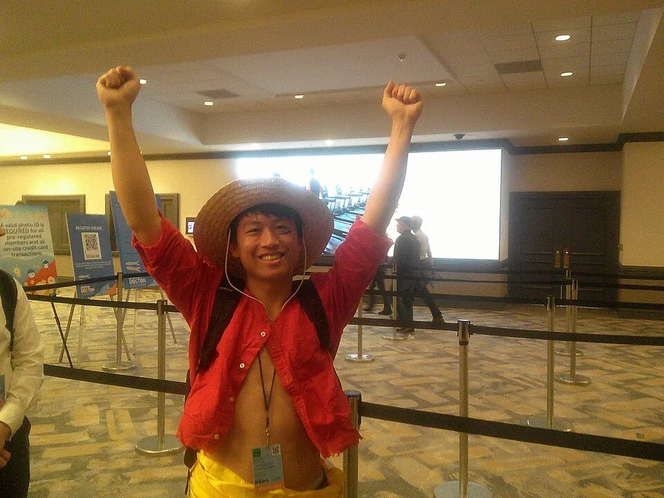 A smiling cosplayer in a straw hat and open red shirt with both fists raised, in a convention hall.