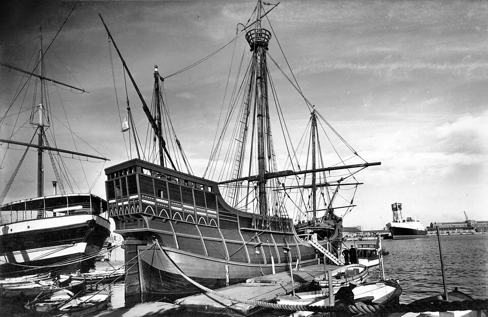 A black-and-white photo of a tall wooden sailing ship with several masts moored in a harbour.