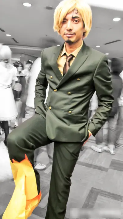 A cosplayer with blond hair swept over one eye and a small goatee, in a dark green suit and tie, with one leg raised in flame-coloured wrapping.