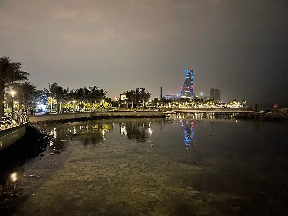 A waterfront promenade at night with palm trees, lamps and a tall glowing tower reflected in calm water.