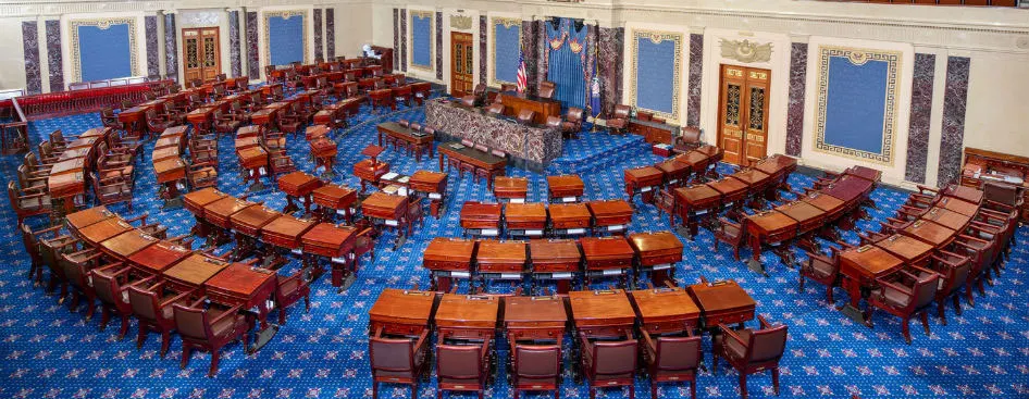 A formal legislative chamber with rows of wooden desks and leather chairs on a blue carpet, facing a raised marble rostrum.