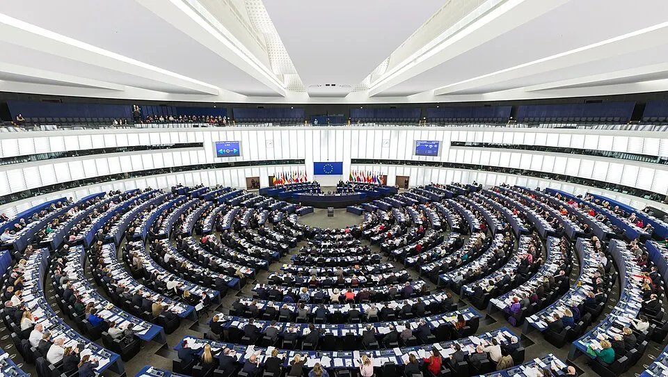 A huge semicircular debating hall filled with rows of desks and members, with flags and a blue banner at the front.