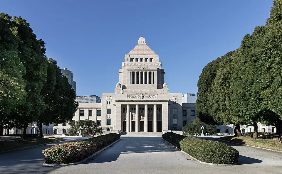 A symmetrical pale stone government building with a stepped pyramid tower, framed by tall trees and clipped hedges.