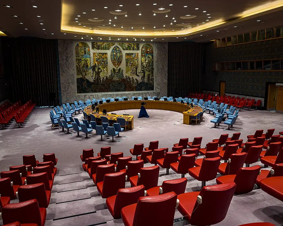An empty council room with a horseshoe-shaped table, blue chairs, red visitor seats and a large mural.