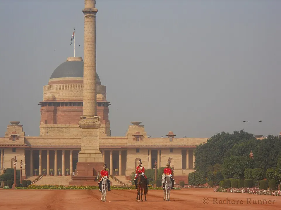 Three red-coated riders on horses on a dusty forecourt in front of a domed sandstone palace and a tall pillar.