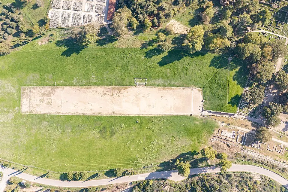 An aerial view looking straight down at a long, flat sandy running track surrounded by grassy slopes and trees.