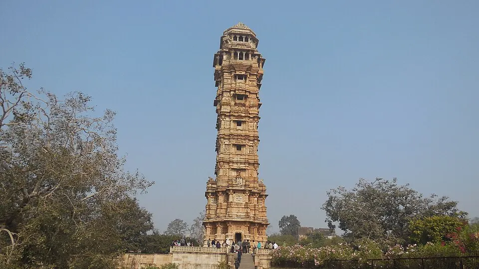 बलुआ पत्थर की बहुमंज़िला ऊँची नक्काशीदार मीनार, जिसके नीचे सीढ़ियाँ और आसपास पेड़ तथा लोग हैं।