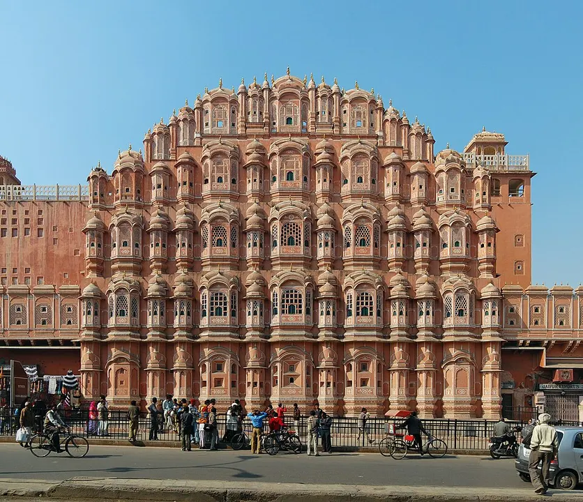 A tall pink sandstone facade covered in rows of small latticed windows and domed balconies stands beside a busy street.