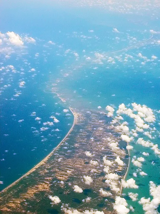 An aerial view of a long, thin curving strip of land and sandbanks stretching across blue-green sea dotted with clouds.