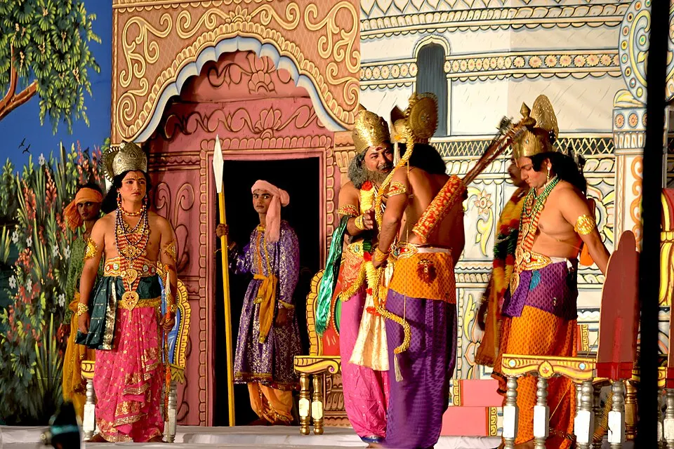 Actors in gold crowns and bright silk costumes on a painted stage set, with a bow and a spear.