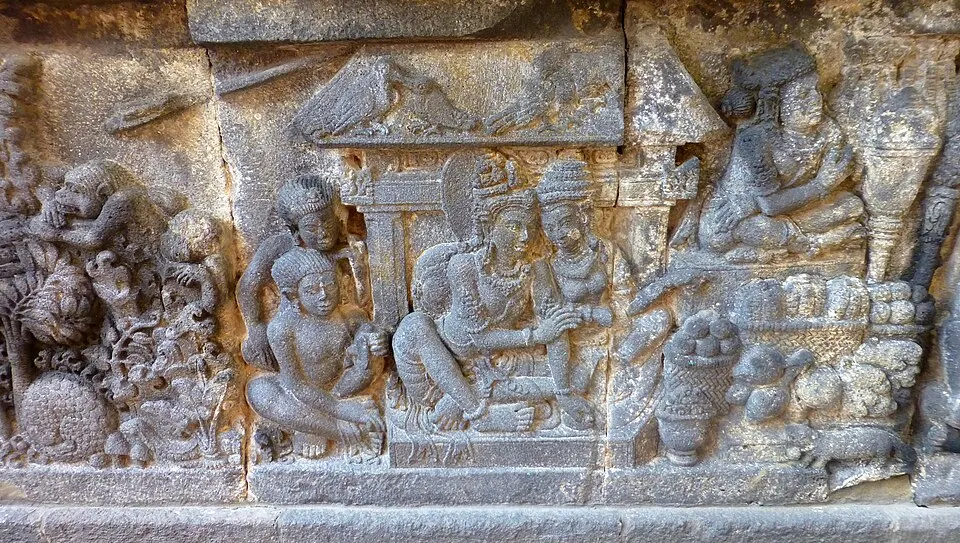 A weathered stone temple relief showing a seated couple under a canopy, with monkeys on the left and fruit baskets on the right.