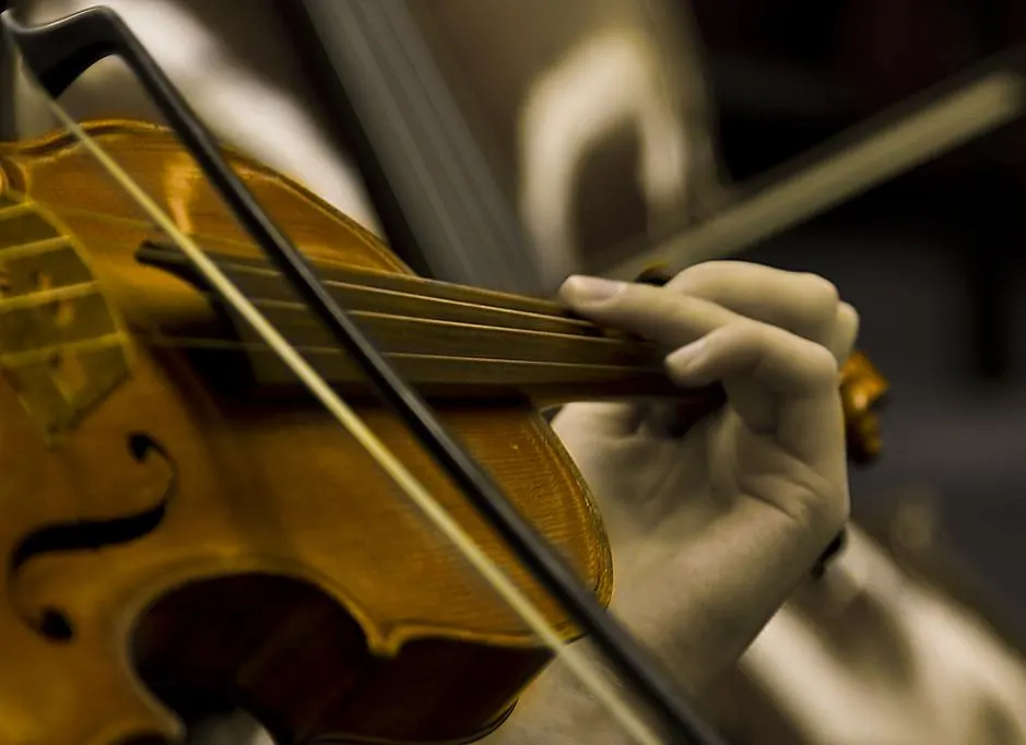 A close-up of a hand pressing the strings of a wooden violin while a bow crosses the frame.