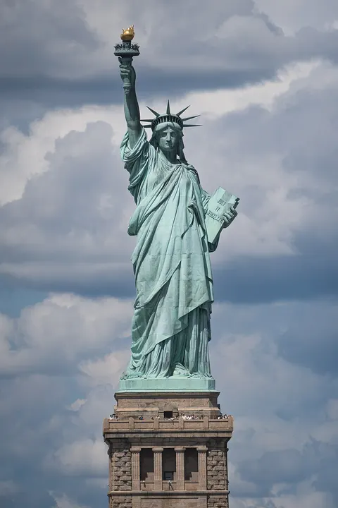 A tall green copper statue of a robed woman holding a torch high and a tablet, wearing a spiked crown, on a stone pedestal against clouds.
