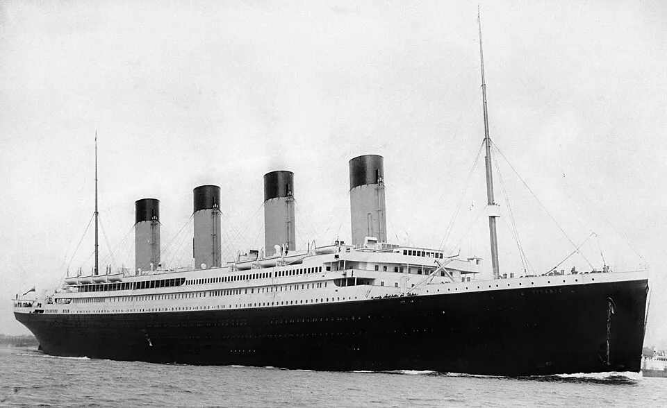 A black and white photograph of a huge ocean liner with four tall funnels and a black hull moving through the water.