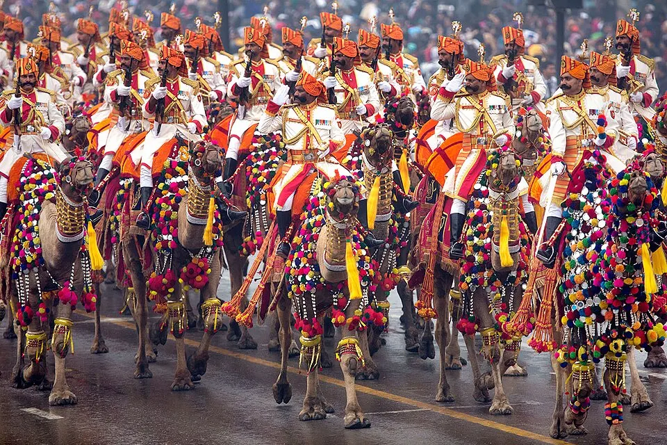 Soldiers in orange turbans and white uniforms riding camels decorated with colourful pom-poms in a parade.