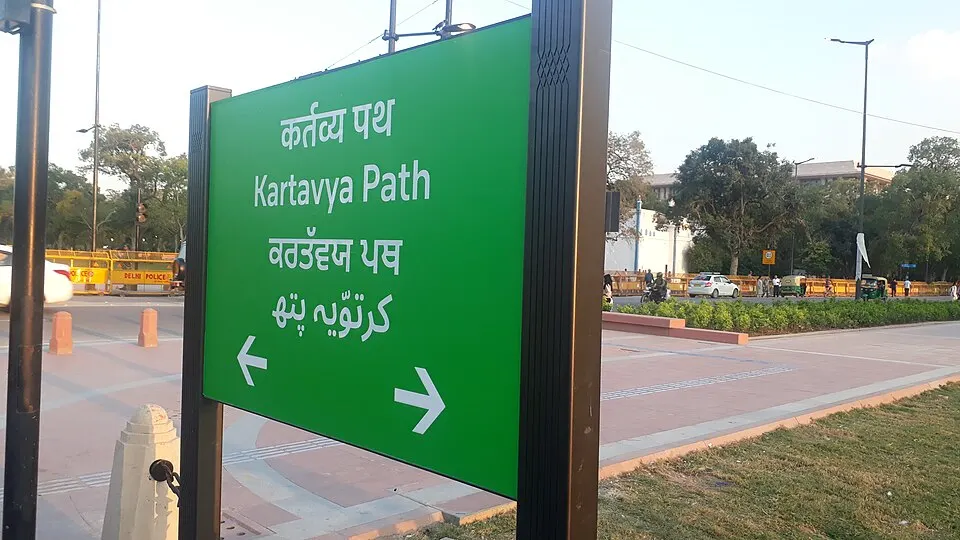 A green road sign in several scripts beside a wide paved avenue with lawns and traffic barriers.