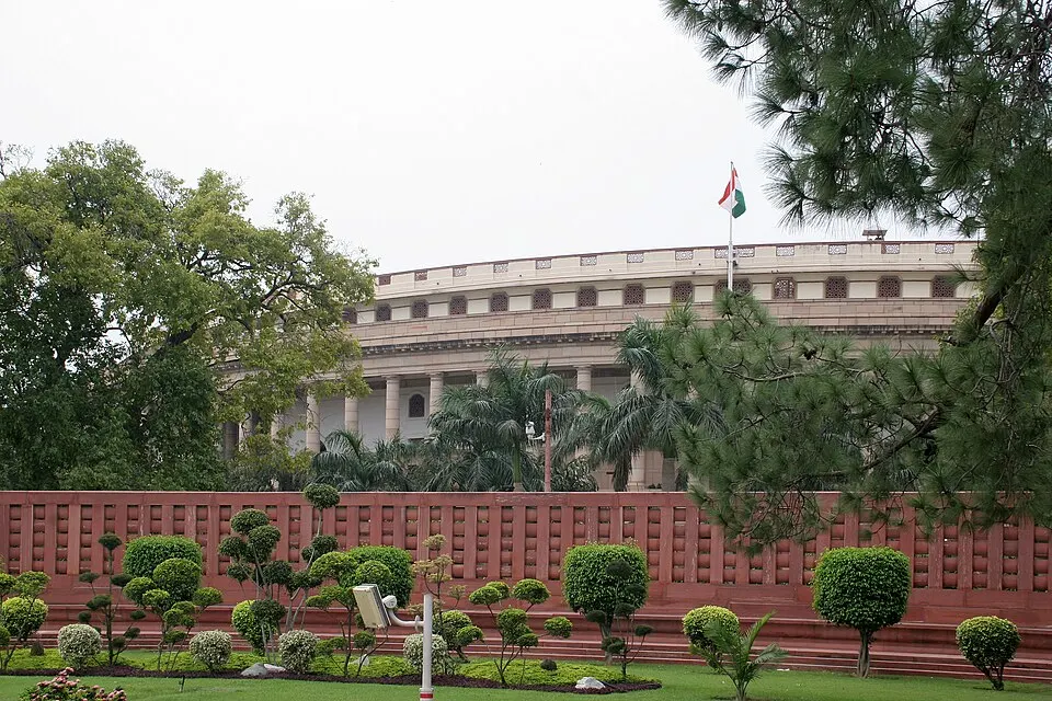 A circular colonnaded stone building behind a red sandstone wall, with trees and a flag on its roof.