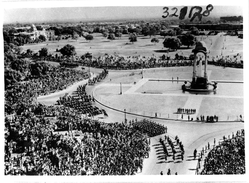 A black and white aerial view of marching columns and huge crowds around a canopied monument on a wide road.
