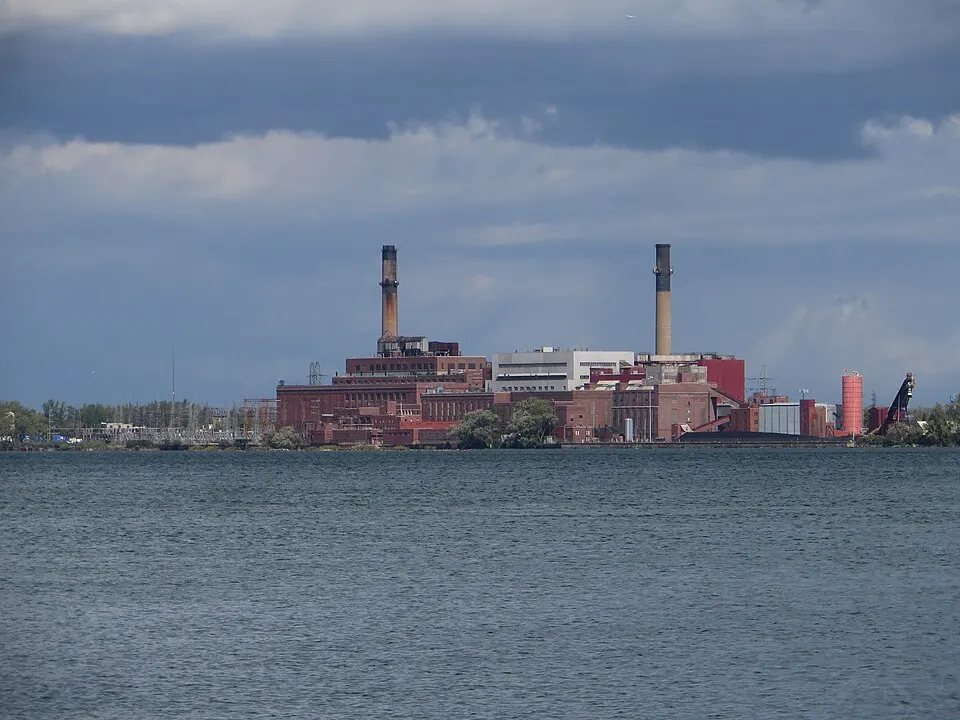 A large brick power station on a lake shore with two tall chimneys under a cloudy sky.