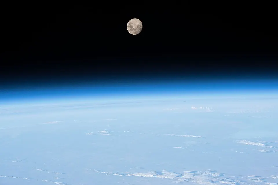 The thin blue glow of Earth's atmosphere along the horizon seen from space, with the Moon above in the black sky.