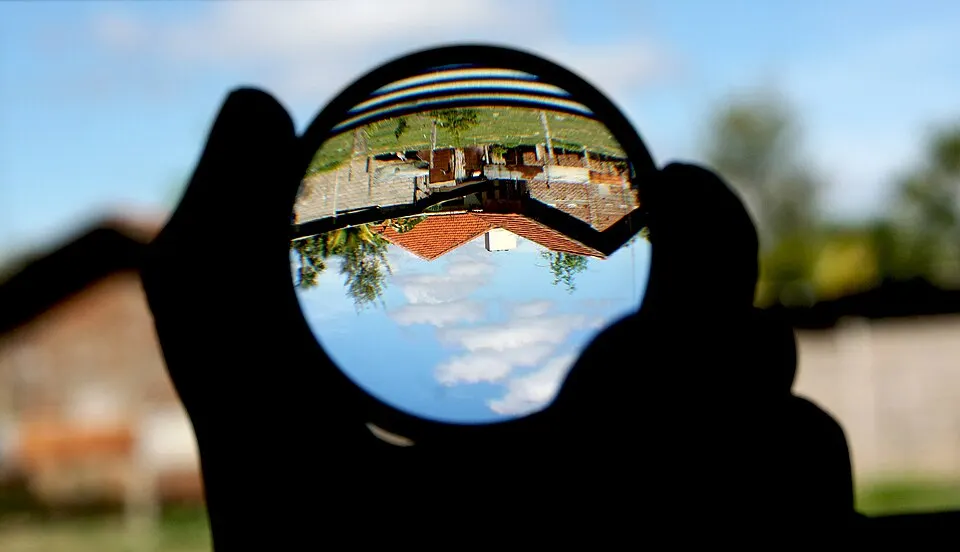 A hand holding a round glass lens that shows an upside-down view of a house and sky.