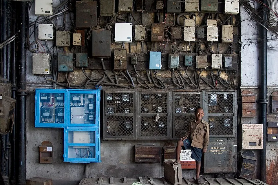 A boy stands beside a wall crowded with old electricity meters, fuse boxes and wires.