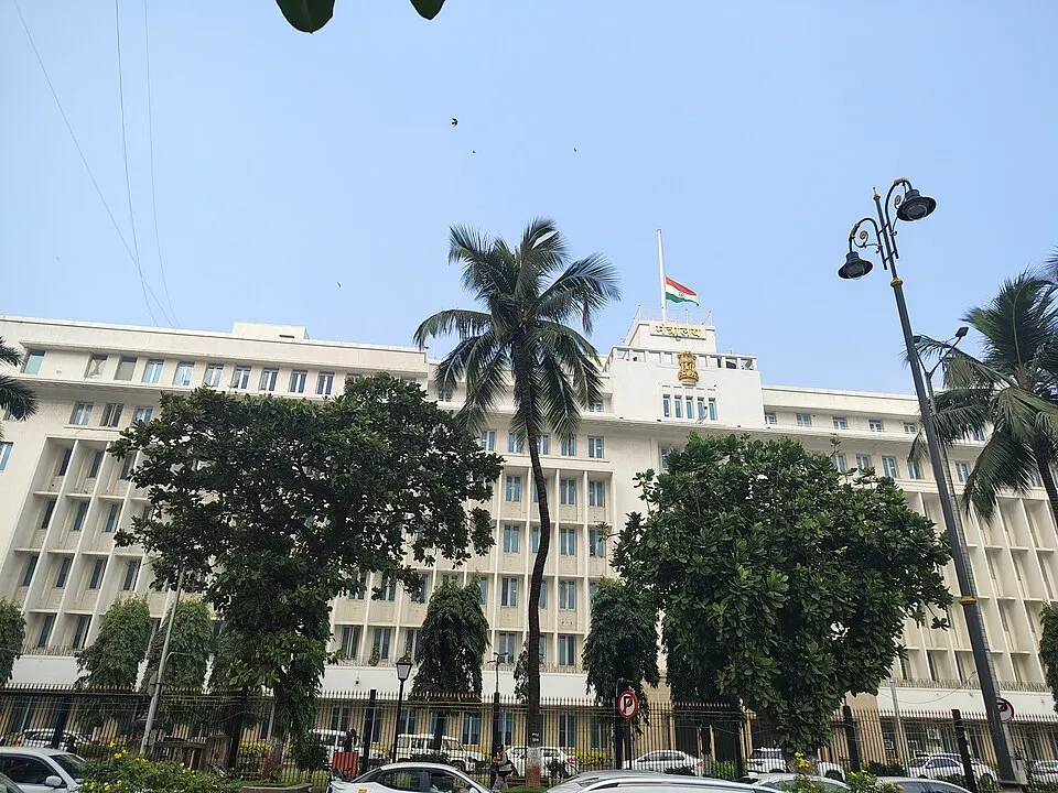 A large cream-coloured government building with a flag on its roof and palm trees in front, behind a black iron fence.