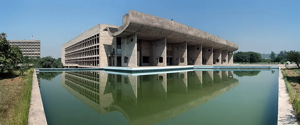 A bare concrete modernist building with a huge curved roof, reflected in a long green pool.