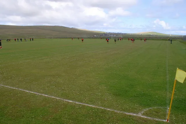 A grassy football pitch with a yellow corner flag and a few players in red far away, with hills behind.