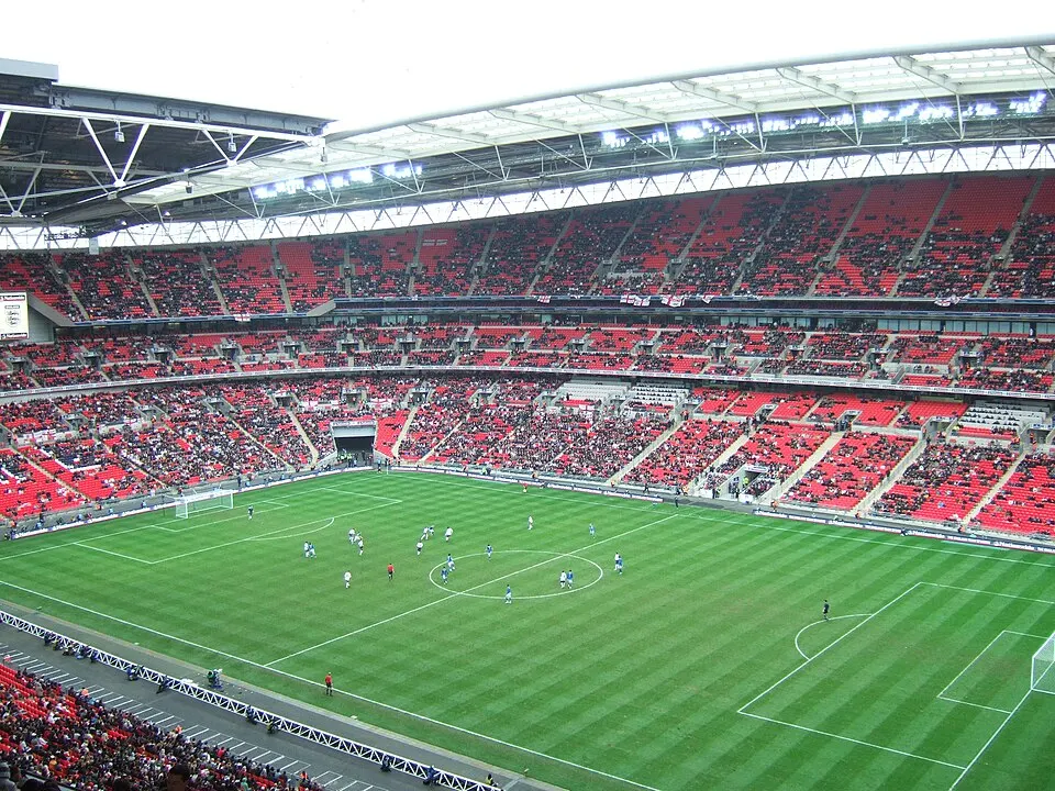 A big stadium with red seats and a floodlit roof, with a football match played far below on the pitch.