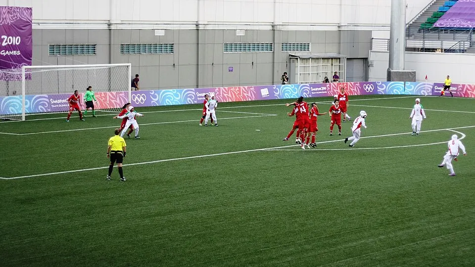 Women in red and white kit chase the ball near a goal on an artificial pitch, with a referee in yellow.