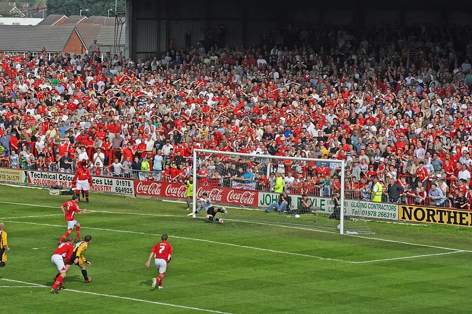 A goalkeeper dives beside the goal as red-shirted players watch, in front of a crowd of fans in red.