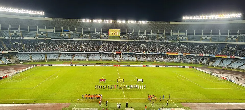 A floodlit football stadium at night with two teams lined up on the pitch in front of a big crowd.