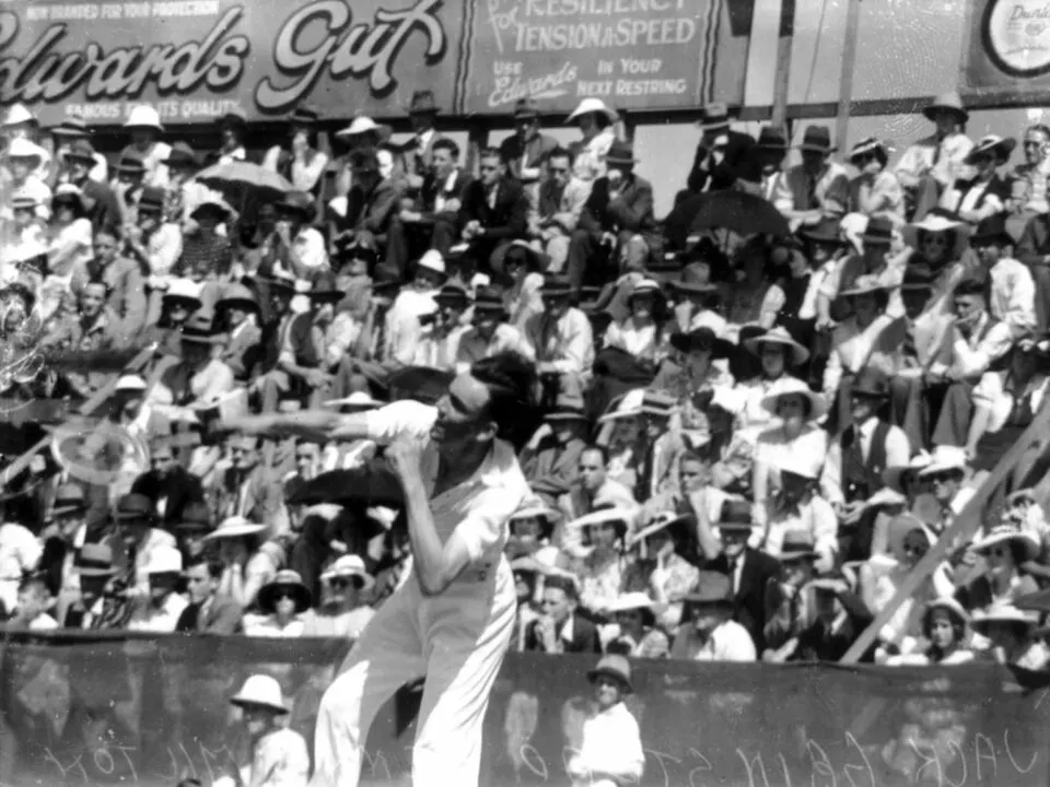 A black-and-white photo of a tennis player in white serving in front of a crowd wearing hats.