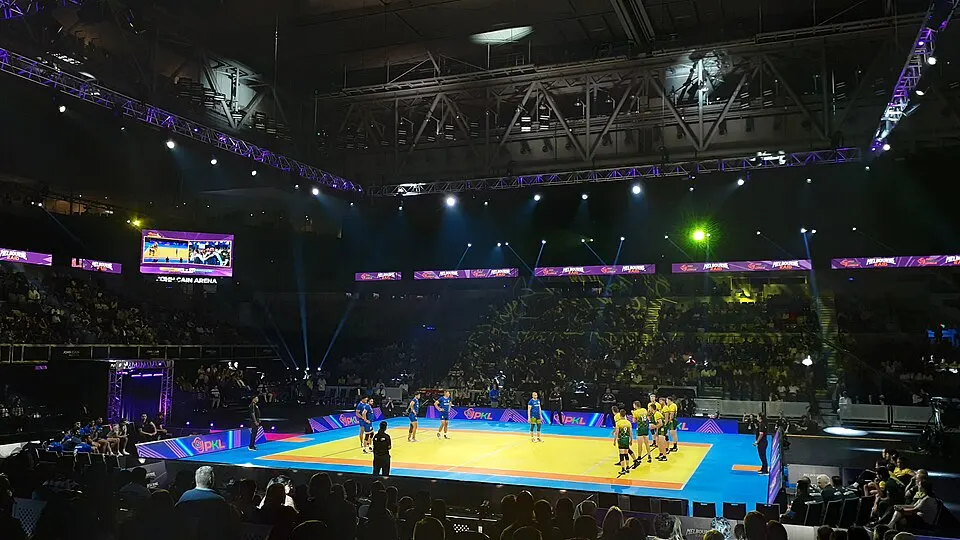 A kabaddi match on a blue and yellow mat inside a dark arena with spotlights and a crowd.