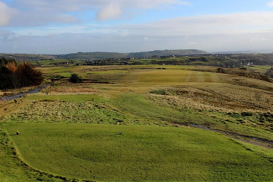 A grassy golf hole viewed from a raised tee, with fairway, a stream and hills in the distance.