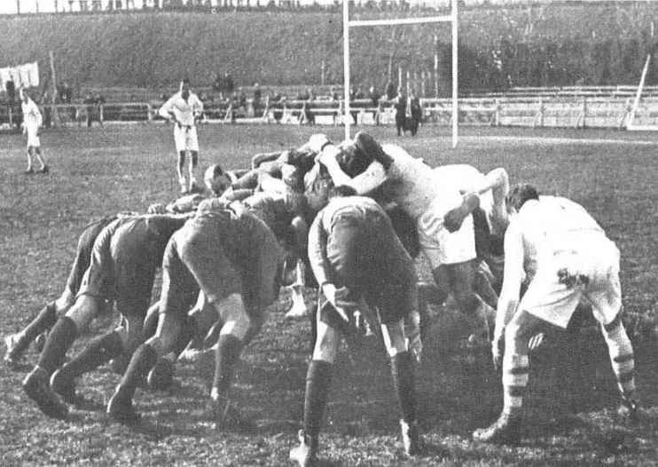 A black-and-white photo of rugby players bunched in a scrum on a field with a goalpost behind.