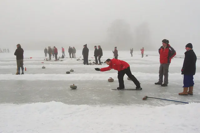 People on a frozen lake slide heavy round stones across the ice, with a person in red lunging forward.