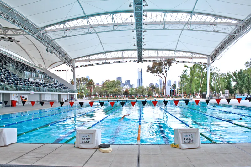 An outdoor swimming pool with lane ropes and a lone swimmer under a large canopy roof.
