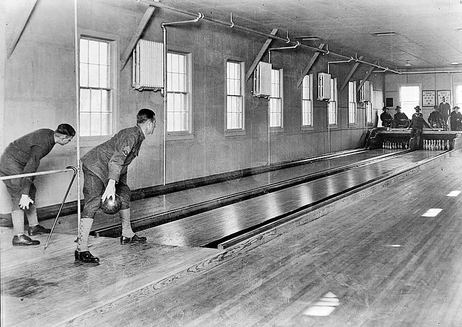 Two men in an old wooden bowling alley, one holding a ball as he lines up a throw down the lane.