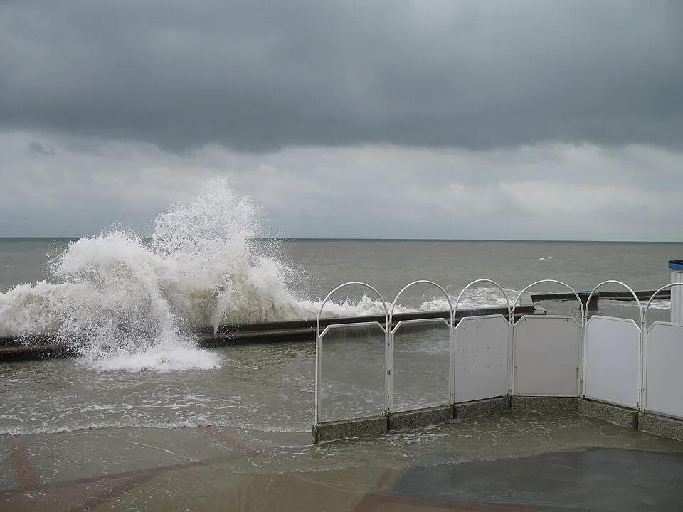 A large wave crashing and splashing against a seafront wall under a heavy grey sky.