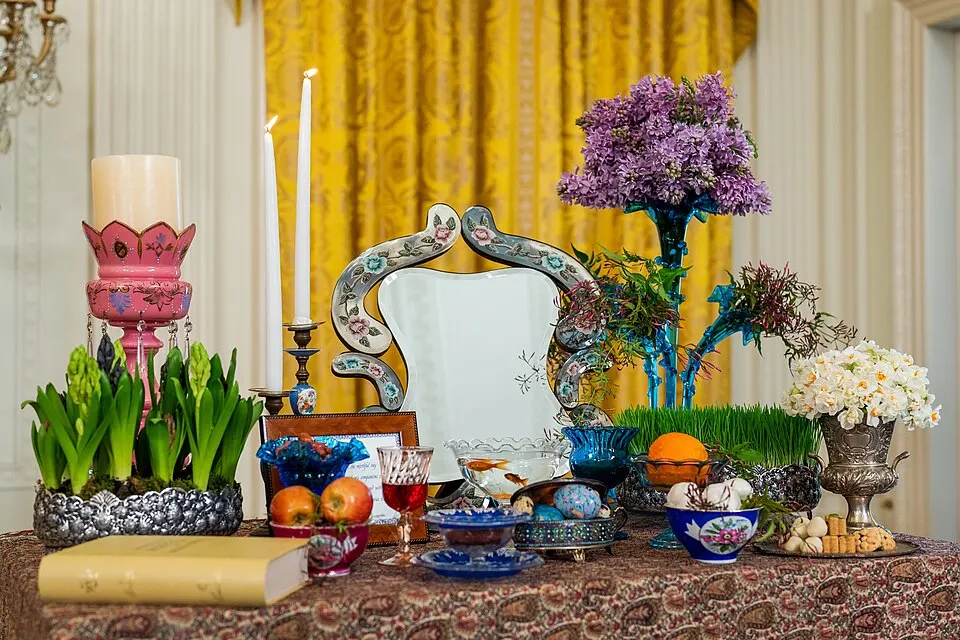 A table set with a mirror, candles, a goldfish bowl, sprouted grass, hyacinths, apples, oranges, painted eggs and a book on a patterned cloth.