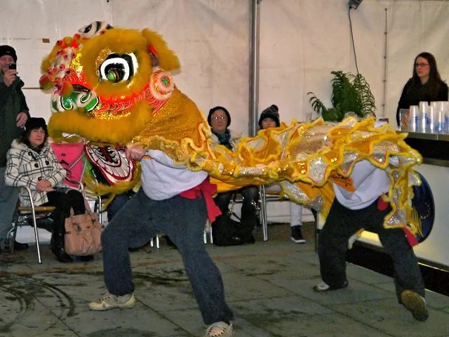 Two people in a bright yellow and gold lion costume performing a dance on a paved floor while people watch.