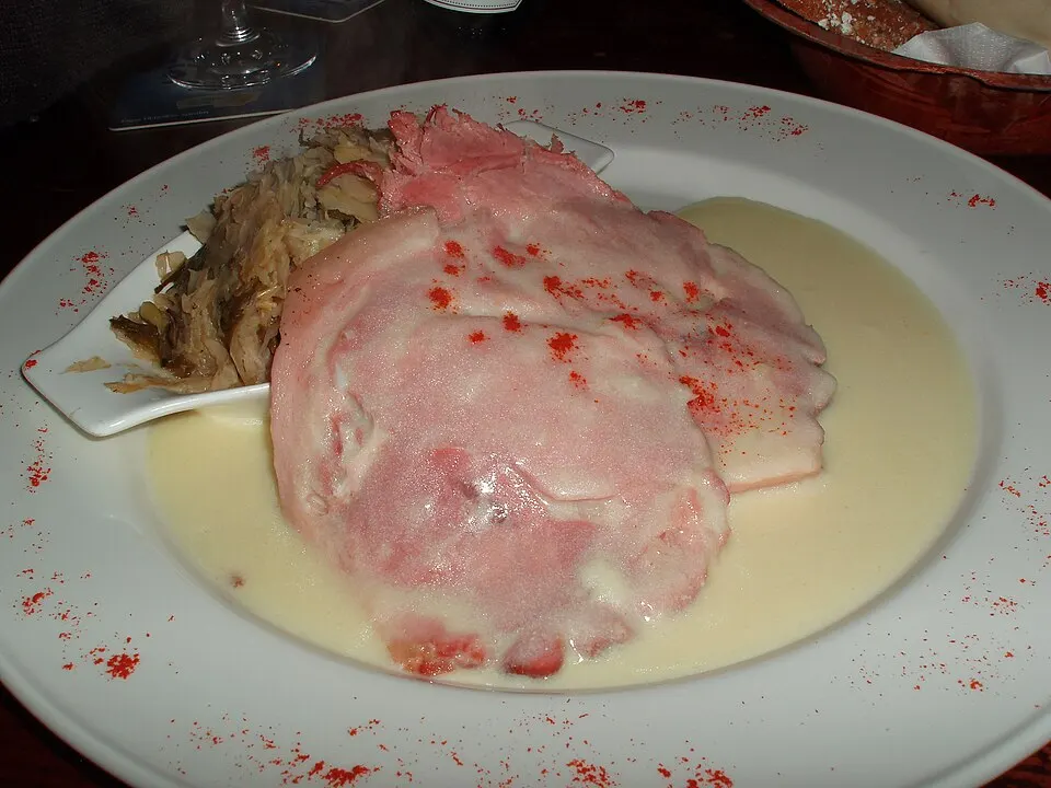 A white plate with slices of pink boiled meat in a creamy sauce, with cooked cabbage on the side.