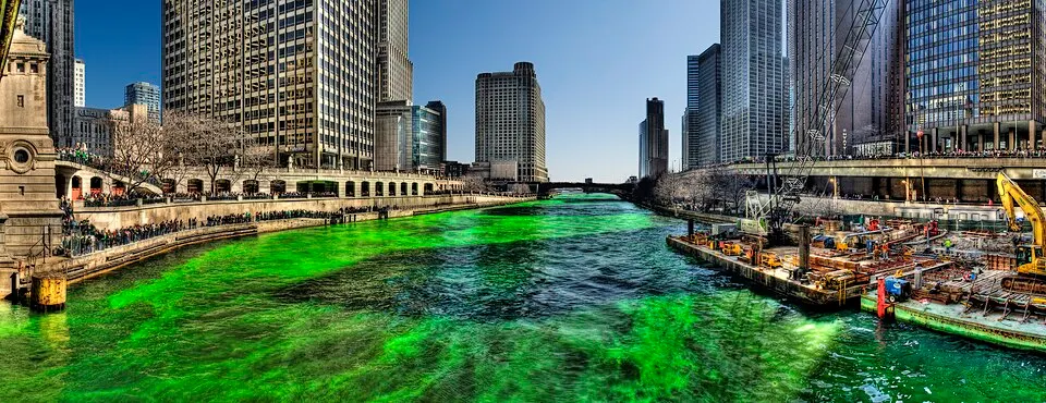 A river running between tall city buildings, coloured bright green, with crowds lining the embankment.