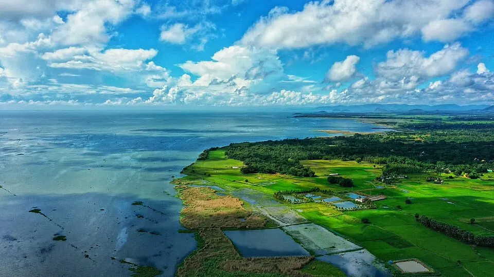 An aerial view of a huge shallow lake meeting green land and fish ponds under a blue sky with big clouds.
