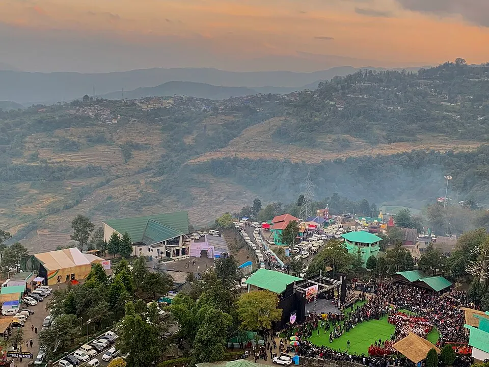 A hillside village venue seen from above at dusk, with a big crowd gathered around an open-air stage and green-roofed huts.
